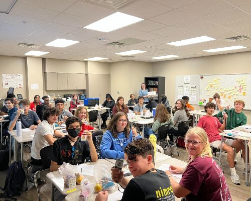  A class of smiling students hold up their IDs on lanyards as the teacher holds boxes of donuts.